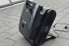 Der Roboter steht senkrecht in seiner schwarzen Ladestation. Sichtbar sind die Lüftungsschlitze an der Seite und das Stromkabel, das am Boden entlang verläuft.