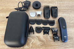 Eine schwarze Transporttasche liegt auf einem Holztisch. Daneben sind ein elektrischer Rasierer von Braun und zahlreiche schwarze sowie silberne Aufsätze und Zubehörteile sauber angeordnet. Alles wirkt ordentlich und vollständig.