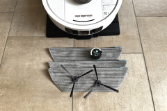 Der Lieferumfang des Shark PowerDetect NeverTouch Pro: Drei Wischpads, zwei Seitenbürsten und eine Duftkapsel neben dem Saugroboter.