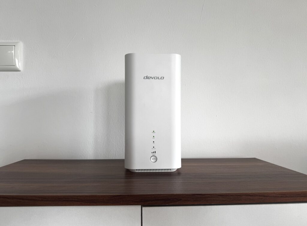 Der devolo-Router auf einem weißen Möbelstück mit dunkler Holzplatte vor einer glatten Wand.